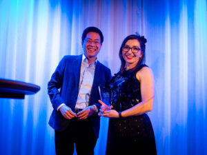 Helen Perris, dressed in black sequins, is presented with her Yamaha Break Award by Managing Director of Yamaha Music Australia, Masa Shibazaki. He is wearing a blue blazer and white shirt and they are standing in front of a lit, blue curtain.
