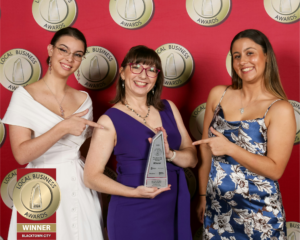 From left to right, Jacki in a white dress, Helen in a purple dress and Bella in a blue and white floral dress stand in front of a red backdrop with the gold local business awards logo repeated across it. Helen is holding a silver trophy and Jacki and Bella are pointing to it. All are smiling.