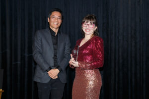 Helen Perris, dressed in a rose and rose-gold sequinned dress, is presented with her Yamaha Break Award by Managing Director of Yamaha Music Australia, Masa Shibazaki. He is wearing a grey blazer and black shirt and they are standing in front of a black curtain.